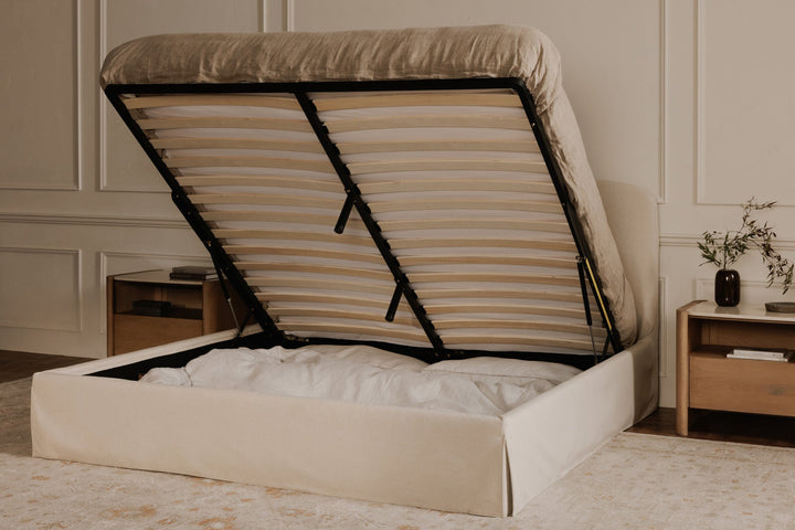 Beige upholstered bed with open storage compartment in a room with wooden nightstands.