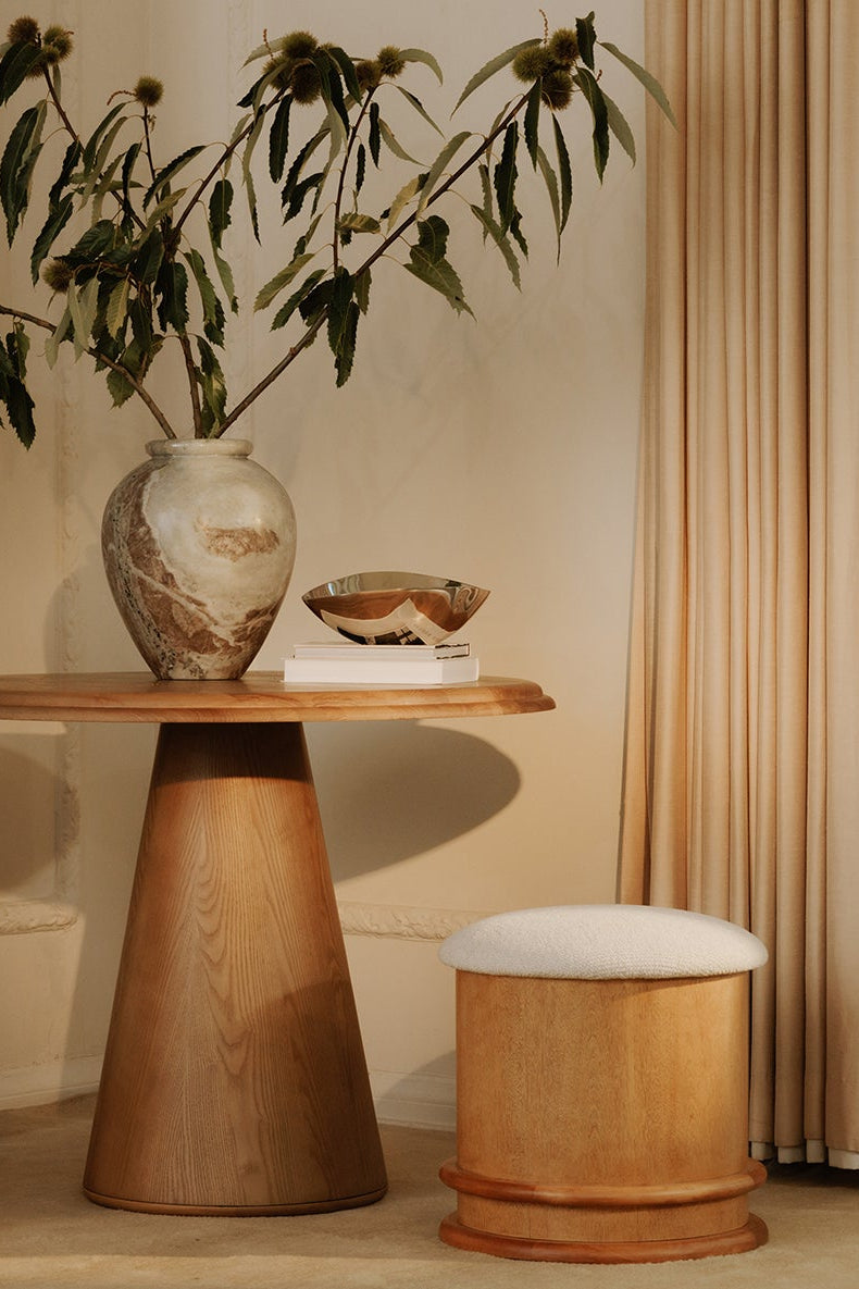 Wooden side table with a vase and plant, next to a wooden stool with a white cushion, against a beige wall.