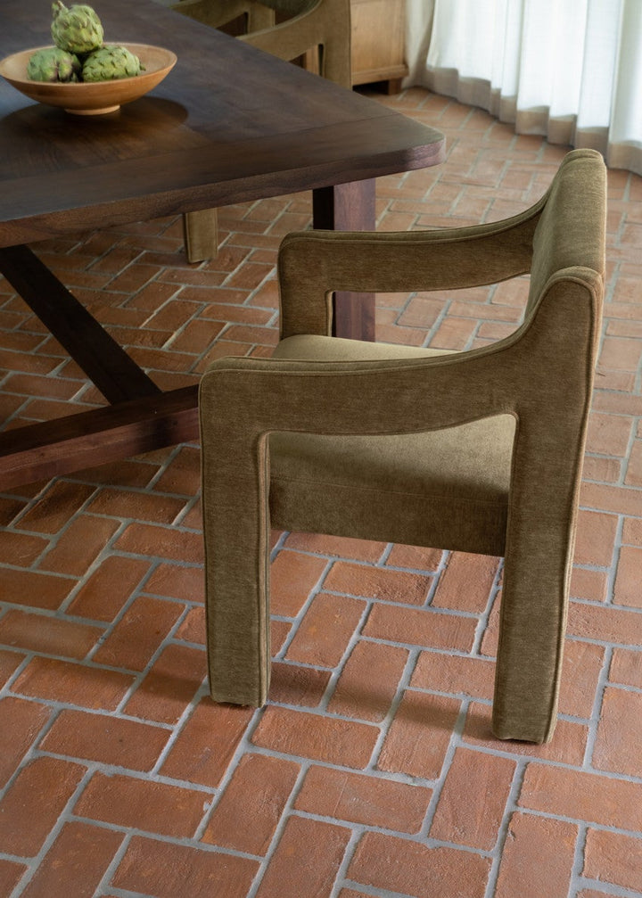 Cedar green upholstered dining chair in a dining area with a wooden table and a bowl of fruit on a brick floor.